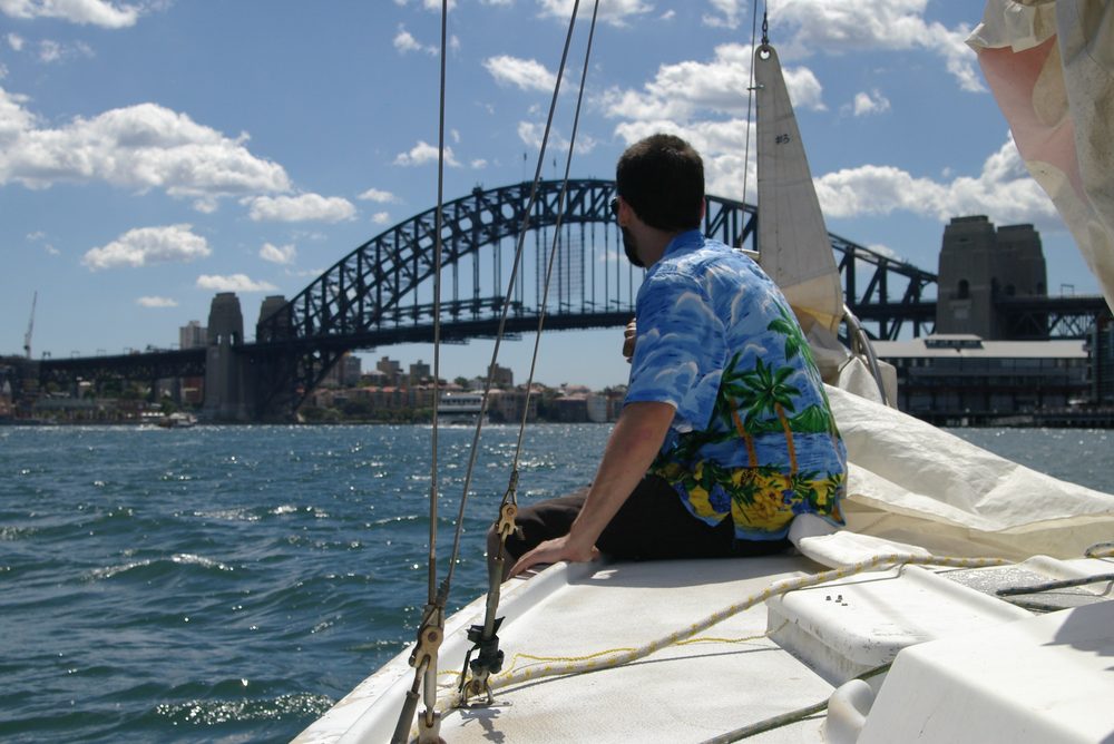 Anthony Fordham in a Hawaiian shirt on the bow of a yacht, Sydney Harbour Bridge ahead