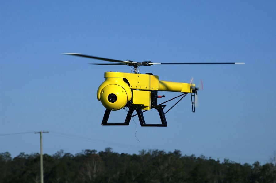 A yellow helicopter drone hovering over gum trees